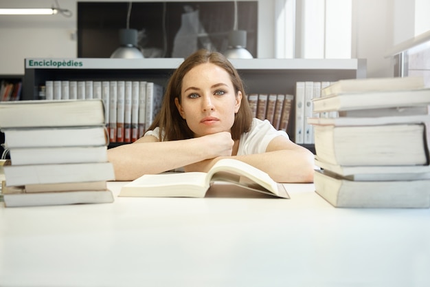 Zelfverzekerde vrouwelijke student studeert aan de bibliotheek, kijkt met een kalme gezichtsuitdrukking aan de tafel met haar kin op haar handen, leert vreemde talen, leest een leerboek