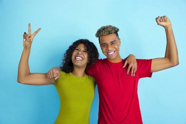 Zich voordoen als winnaars, lachend. Jonge emotionele Afro-Amerikaanse man en vrouw in kleurrijke kleding op blauwe muur.