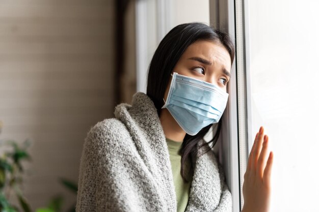 Ziek Aziatisch meisje met medisch gezichtsmasker dat bij het raam staat en ernaar verlangt om naar buiten te gaan terwijl ze op de...