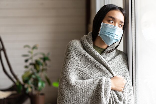 Ziek Aziatisch meisje met medisch gezichtsmasker dat bij het raam staat en ernaar verlangt om naar buiten te gaan terwijl ze op...