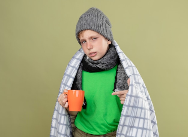 Ziek jongetje draagt groen t-shirt in warme sjaal en muts gewikkeld in een deken met kopje hete thee wijzend met vinger erop staande over lichte muur