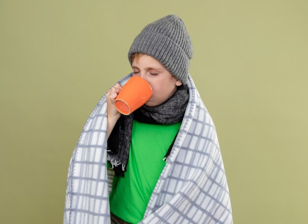 Ziek jongetje dragen groene t-shirt in warme sjaal en muts gewikkeld in een deken hete thee drinken staande over lichte muur