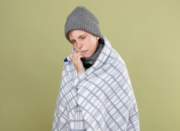 Ziek jongetje groen t-shirt dragen in warme sjaal en muts gewikkeld in deken thermometer aanbrengend mond meten temperatuur staande boven lichte muur