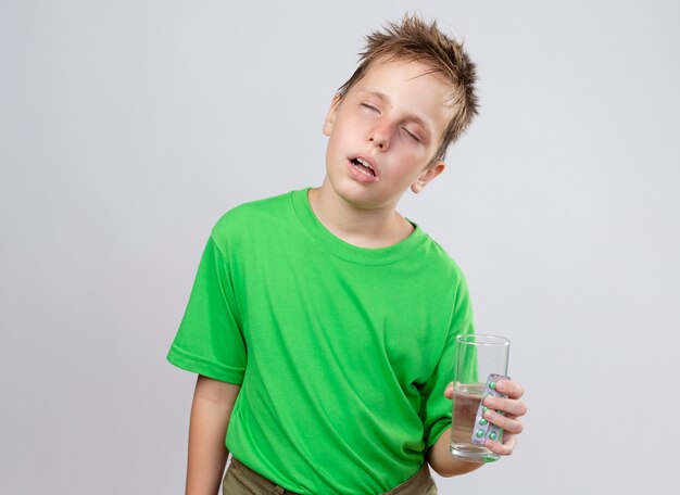 Ziek jongetje in groen t-shirt die zich onwel voelt met glas water en pillen die aan koude lijden die zich over witte muur bevinden