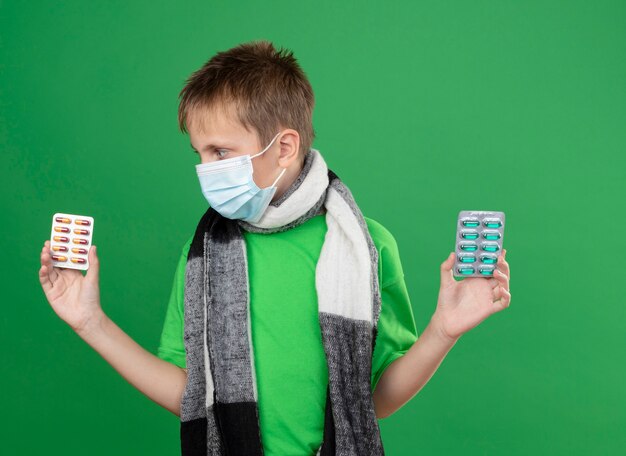 Ziek jongetje in groen t-shirt en warme sjaal rond zijn versleten gezicht beschermend masker met pillen op zoek verward proberen om een keuze te maken staande op groene achtergrond