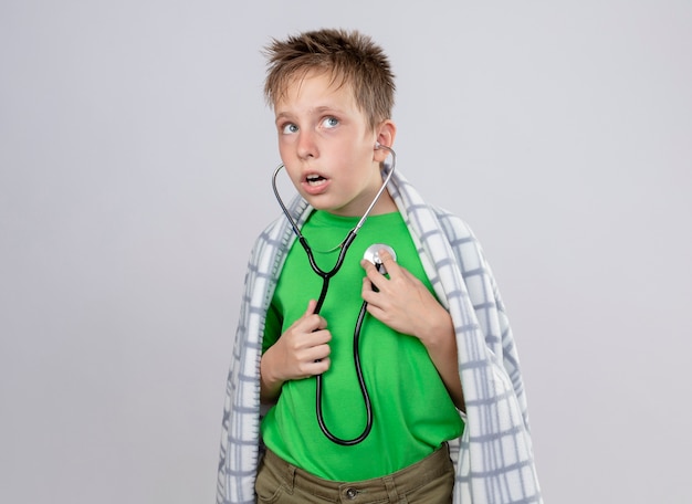 Ziek jongetje in groen t-shirt gewikkeld in deken onwel voelen met een stethoscoop om zijn nek luisteren naar de hartslag die verward is staande over een witte muur
