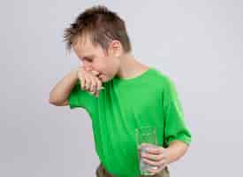 Gratis foto ziek jongetje in groen t-shirt zich onwel voelen met glas water en pillen neus afvegen met papieren servet staande over witte muur