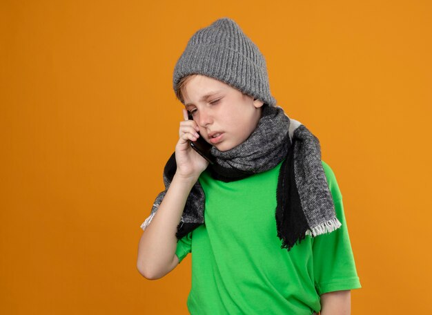 Ziek jongetje met groen t-shirt in warme sjaal en muts die zich onwel voelen praten over de mobiele telefoon die zich over de oranje muur bevindt