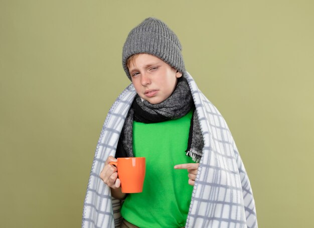 Ziek jongetje met groen t-shirt in warme sjaal en muts gewikkeld in een deken met kopje hete thee wijzend met de vinger erop ongelukkig en ziek staande over lichte muur