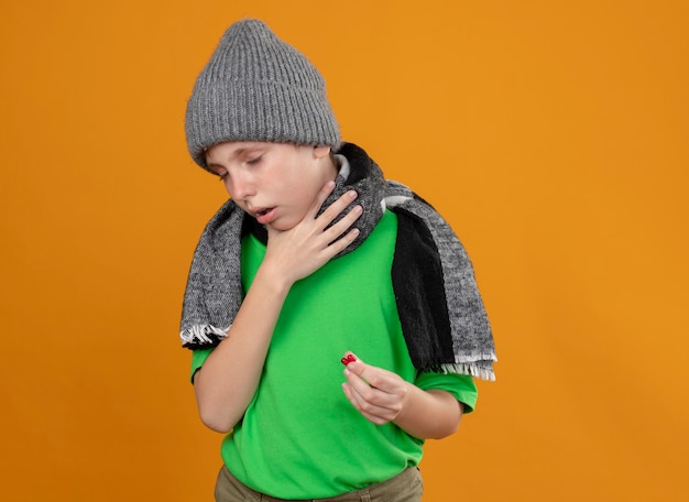 Ziek jongetje met groen t-shirt in warme sjaal en muts met pillen die zich onwel voelen ziek en ongelukkig hoesten staande over oranje muur