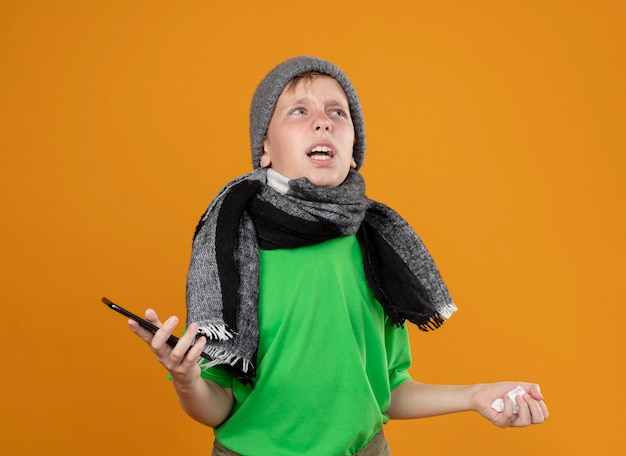 Ziek jongetje met groen t-shirt in warme sjaal en muts zich onwel voelen met smartphone en papieren servet opzij kijken verward en ontevreden over oranje muur staan