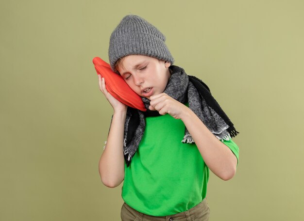 Ziek jongetje met groen t-shirt in warme sjaal en muts zich onwel voelen met waterfles om warm te blijven, leunend hoofd erop hoesten staande over lichte muur