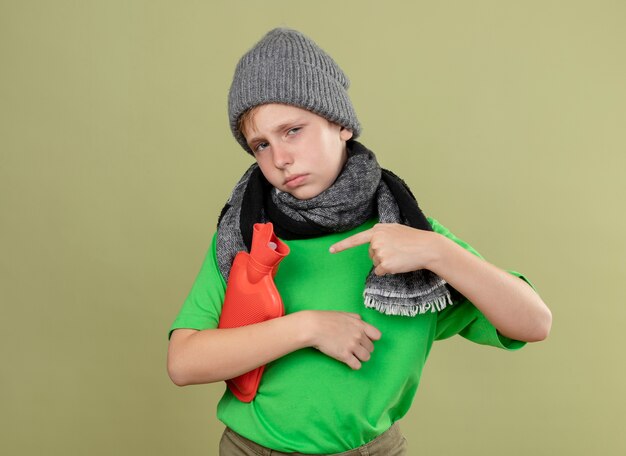 Ziek jongetje met groen t-shirt in warme sjaal en muts zich onwel voelen met waterfles om warm te blijven, wijzend met de vinger erop staande over lichte muur