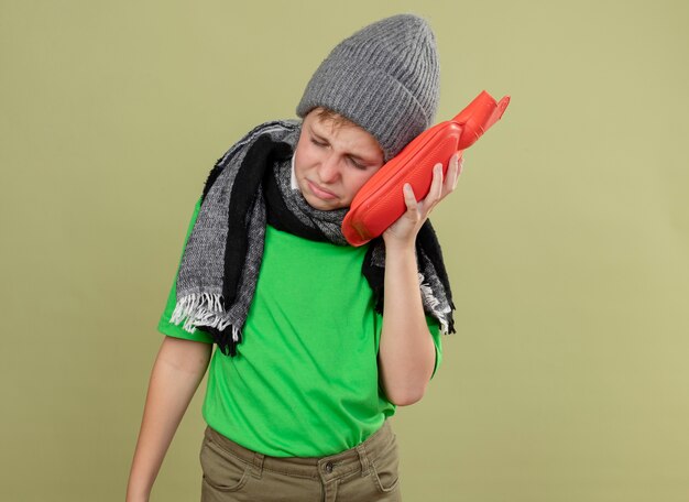 Ziek jongetje met groen t-shirt in warme sjaal en muts zich onwel voelen met waterfles om warm te houden, leunend hoofd erop, ziek en ongelukkig staande over lichte muur