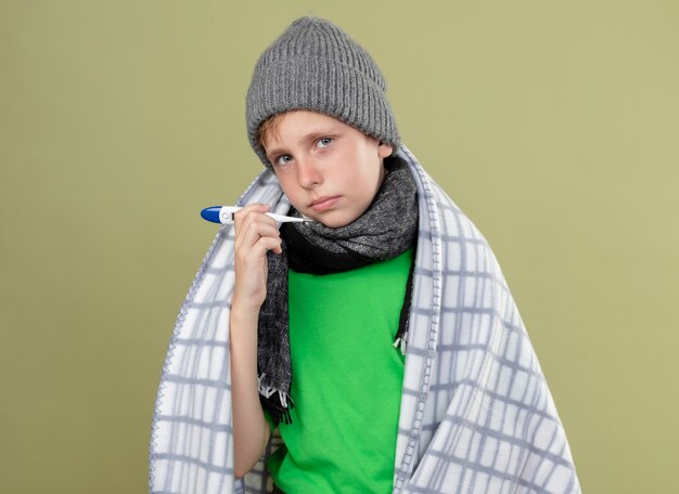 Ziek jongetje met groene t-shirt in warme sjaal en muts gewikkeld in een deken met thermometer ongelukkig en ziek staande over lichte muur