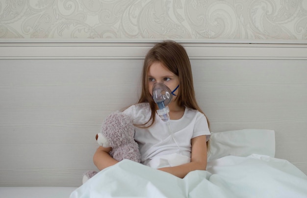 Ziek kind is ziek thuis, liggend in bed met teddybeer. voert de inhalatieprocedure uit met een vernevelaar van de luchtwegen. behandeling van een kind voor hoest. compressie inhalator. selectieve aandacht.
