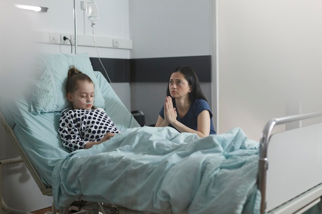 Gratis foto ziek kind onder behandeling slaapt in de kinderafdeling van het ziekenhuis terwijl een bezorgde moeder naast haar zit. ziek meisje rust op het bed van de patiënt terwijl moeder naast haar bidt
