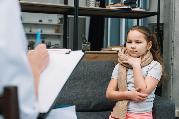 Ziek meisje met sjaal rond haar halszitting voor vrouw die op klembord met pen schrijven