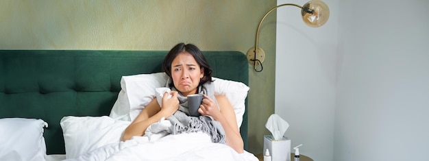 Ziek, verdrietig aziatisch meisje dat in haar bed ligt met griep, wordt verkouden, neemt pillen en drinkt hete theegevoel