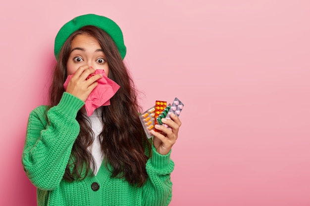 Zieke Aziatische vrouw verkouden tijdens natte herfstdag, heeft lopende neus, houdt verschillende pillen voor het genezen van ziekte, draagt groene baret en trui