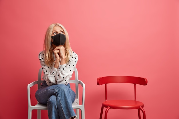 Zieke blonde vrouw houdt handen onder de kin en kijkt bedachtzaam ergens draagt beschermend masker tegen coronavirus herstelt van ziekte thuis zit op stoel binnen