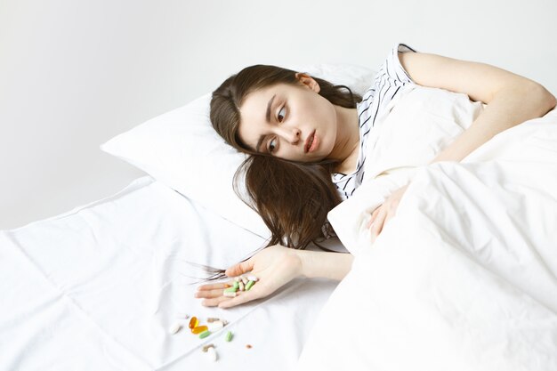 zieke jonge brunette vrouw ochtend doorbrengen in bed, stelletje pillen op haar hand kijken en gemorst over wit vel