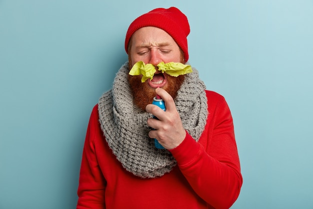 Zieke man in warme kleren met papieren zakdoekjes in de neus en spray voor keelpijn