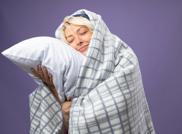 Zieke ongezonde vrouw met kort haar in warme muts gewikkeld in een deken met kussen haar hoofd leunend op kussen met gesloten ogen glimlachend over paarse achtergrond