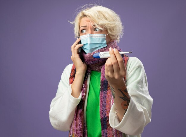 Zieke ongezonde vrouw met kort haar in warme sjaal en beschermend gezichtsmasker met thermometer op zoek bezorgd tijdens het praten op mobiele telefoon, staande over paarse muur