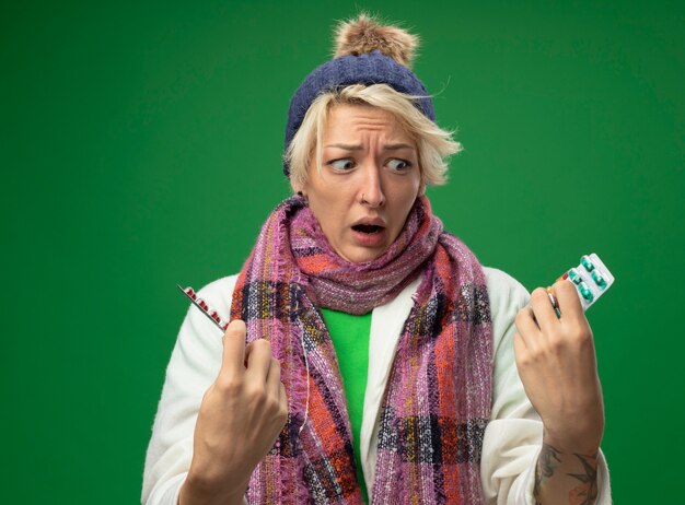 Zieke ongezonde vrouw met kort haar in warme sjaal en muts zich onwel voelen met blaren met pillen kijken camera bezorgd en verward proberen te maken keuze staande op groene achtergrond