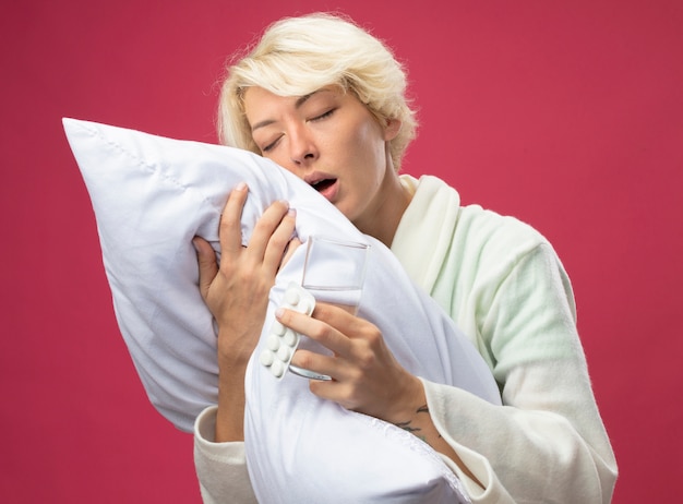 Zieke ongezonde vrouw met kort haar knuffelen kussen onwel gevoel met glas water en medicijnen staande over roze muur