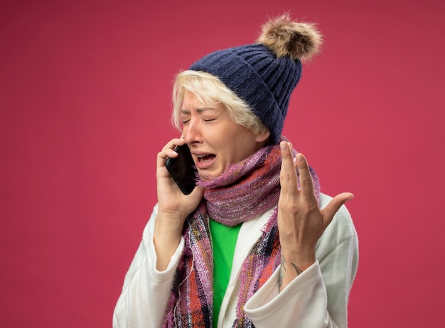 Zieke, ongezonde vrouw van streek met kort haar in warme sjaal en hoed, zich onwel voelen, huilen tijdens het praten op mobiele telefoon met opgeheven arm staande over roze muur