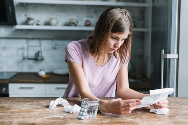 Gratis foto zieke vrouw die voorschrift met blaarpak en glas water op houten bureau bekijkt