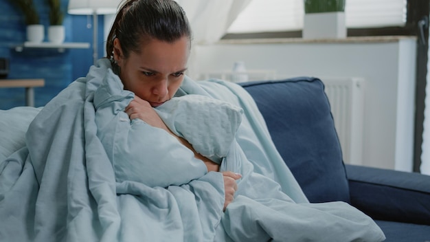 Zieke vrouw met griep die deken en kussen gebruikt tegen thuis rillen. Zieke volwassene die het koud heeft en koude rillingen heeft vanwege koortssymptomen. Close up van persoon met ziekte zittend op de bank