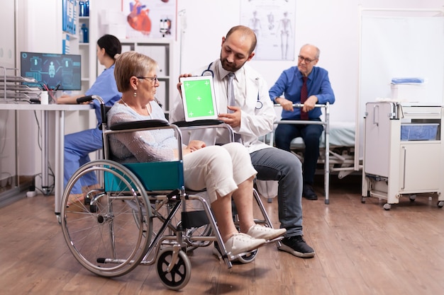 Ziekenhuispersoneel raadpleegt gehandicapte oudere vrouw die in een rolstoel zit en tablet-pc vasthoudt met groen scherm