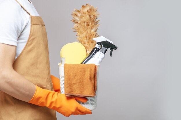 Zijaanzicht, close-up hand van jonge man in schort en rubberen handschoenen met een mand met reinigingsapparatuur, de plumeau, spuitfles, spons en doek om in de mand af te vegen