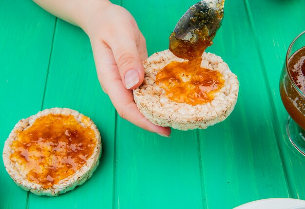 Zijaanzicht dat van hand knapperig knäckebrood met perzikjam erop en lepel op groene oppervlakte houdt