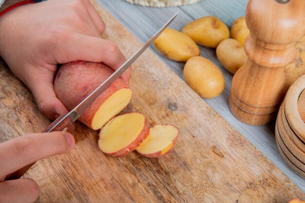 Zijaanzicht die van handen rode aardappel met mes op scherpe raad snijden en zout met nieuwe aardappels op houten lijst