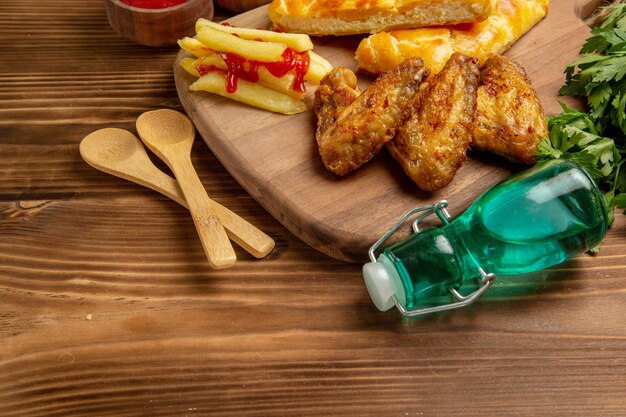 Zijaanzicht fastfood aan boord frietjes met ketchup twee stukken taart kippenvleugels op het keukenbord naast de kom ketchup houten lepels kruiden en fles