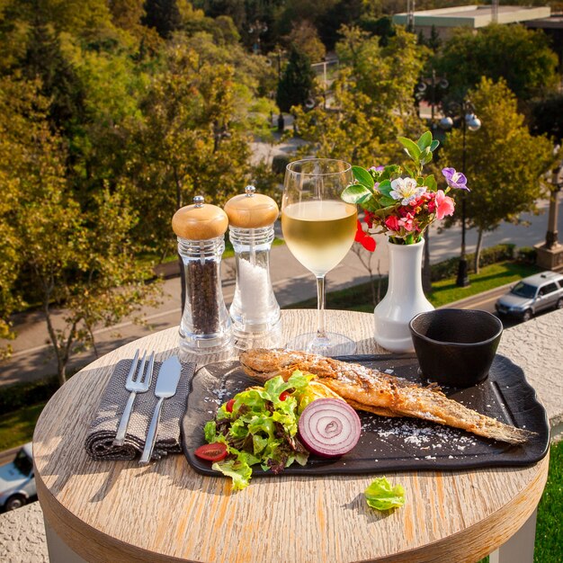 Zijaanzicht gebakken vis met sla, tomaten, uien en saus op een zwarte plaat, een glas witte wijn op een kleine ronde tafel met uitzicht op de stad