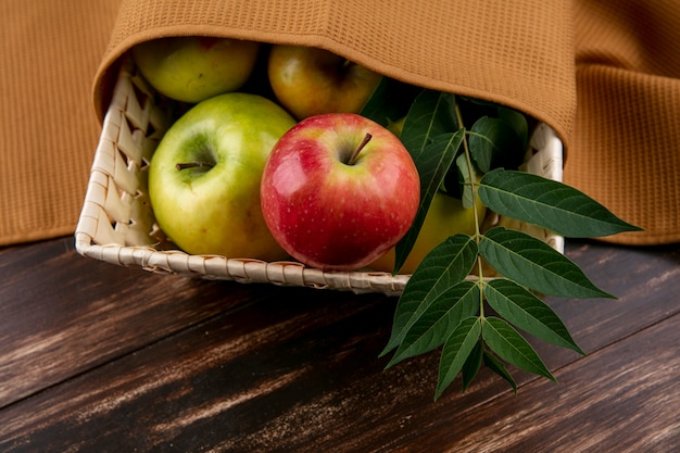 Zijaanzicht groene en rode appels in een mand met een tak en een bruine handdoek op een houten achtergrond