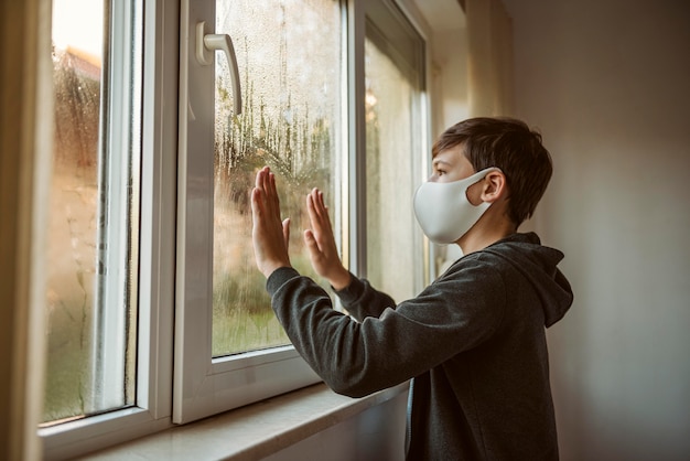 Zijaanzicht kleine jongen met gezichtsmasker kijkt door het raam