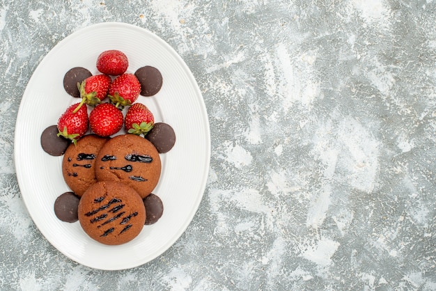 Zijaanzicht linksboven chocoladekoekjes aardbeien en ronde chocolaatjes op het witte ovale bord op de grijswitte ondergrond