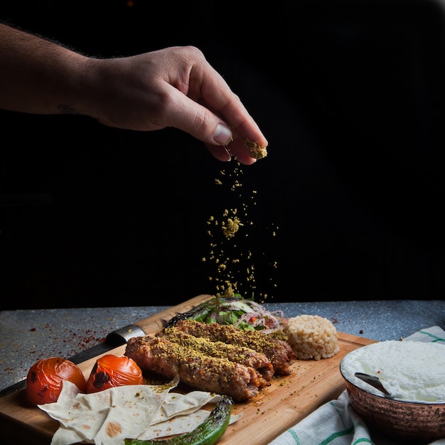 Zijaanzicht lule kebab met tomaat en papier en ayran en hand voegt kruiden toe in serveerplank