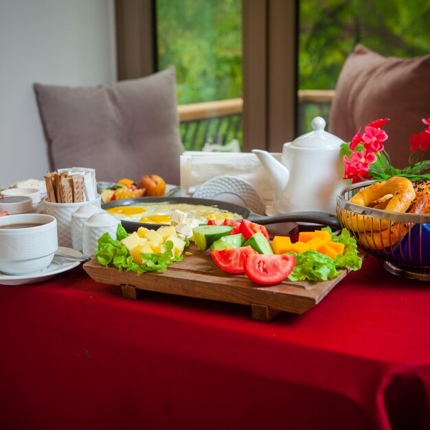 Zijaanzicht ontbijt op een tafel met een rood tafellaken gebakken eieren, kaas, kaas, komkommers, tomaten, sla, koffie