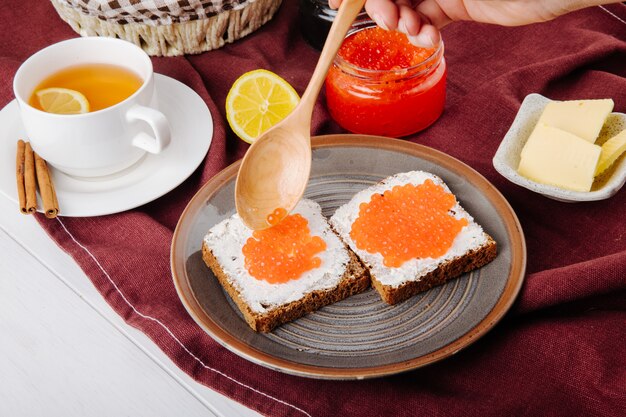 Zijaanzicht rode kaviaar toast roggebrood met cottage cheese rode kaviaar boter kopje thee en schijfje citroen op tafel