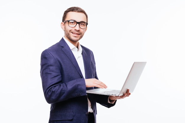 Zijaanzicht van aantrekkelijke jonge zakenman in klassiek pak met behulp van laptop, staande tegen een witte muur