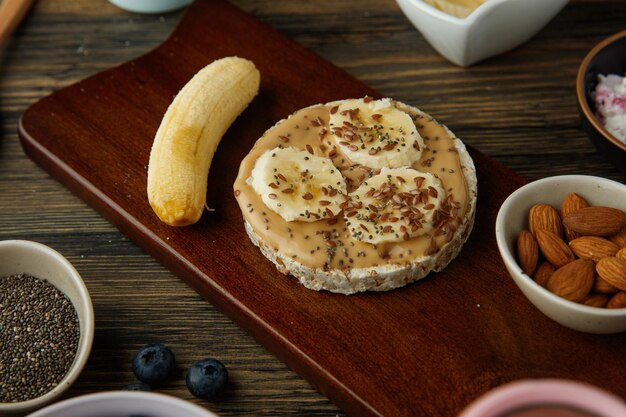 Zijaanzicht van banaan-pindakaas-cripbread-snack met banaan en amandelen op snijplank en sleedoorn op houten ondergrond