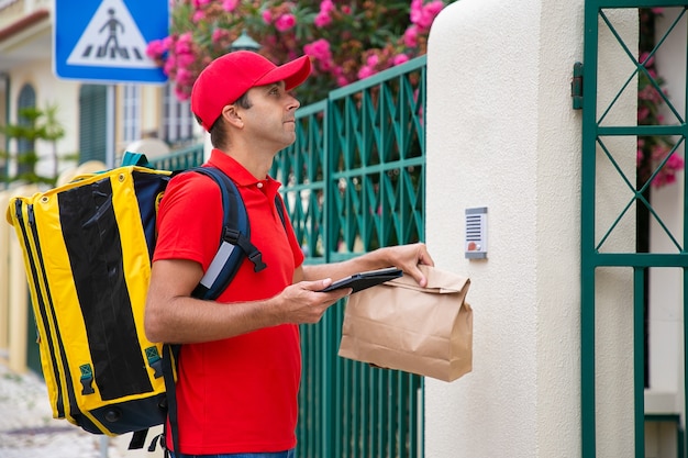 Zijaanzicht van bezorger die in rood GLB op klant wacht bij ingang. Serieuze koerier met gele thermische rugzak en pakket dat de expresbestelling aan de klant levert. Bezorgservice en postconcept