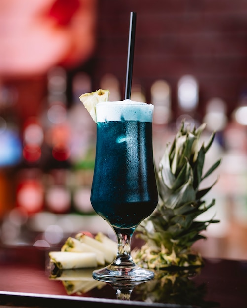 Zijaanzicht van blauwe curacao cocktail in een hoog glas met ananas segment op tafel